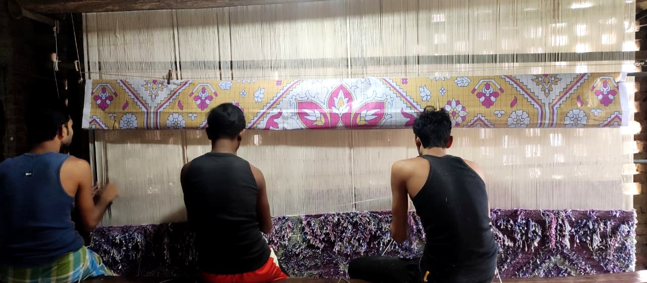 Stage 01 — the design drawing hung above the loom, three weavers reading it row by row