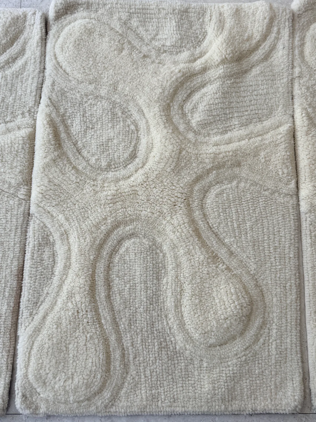 Close view of a carved bench-tufted rug in undyed cream, showing raised curved relief shapes against a lower pile ground