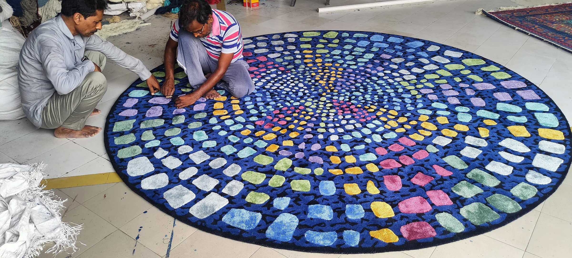 A colourful mosaic-pattern round rug being carved by two finishers to raise motifs against the ground