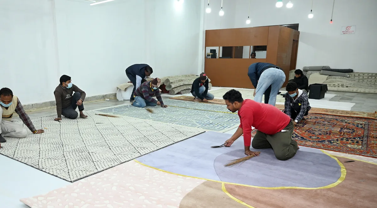 Wide view of the PiHue finishing floor in Bhadohi &mdash; rugs on inspection benches, checked and measured before packing