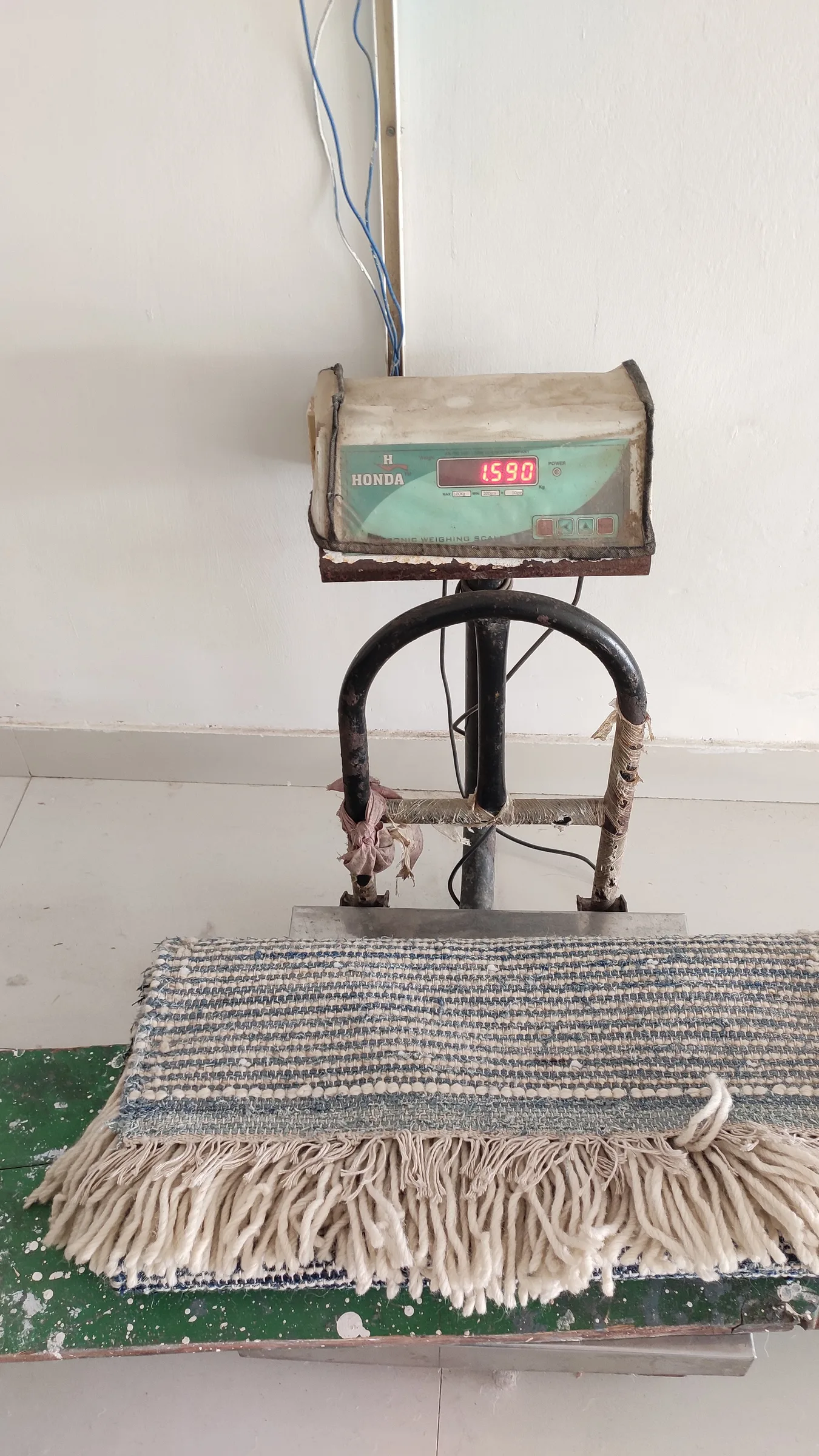 A finished flatweave rug suspended on a hanging scale — digital display reading 1590 grams