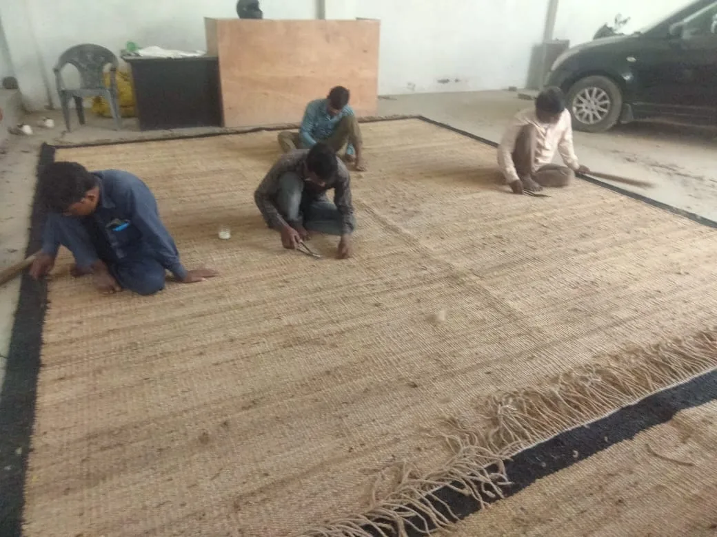Four finishers hand-binding the edge of a large jute rug on a warehouse floor