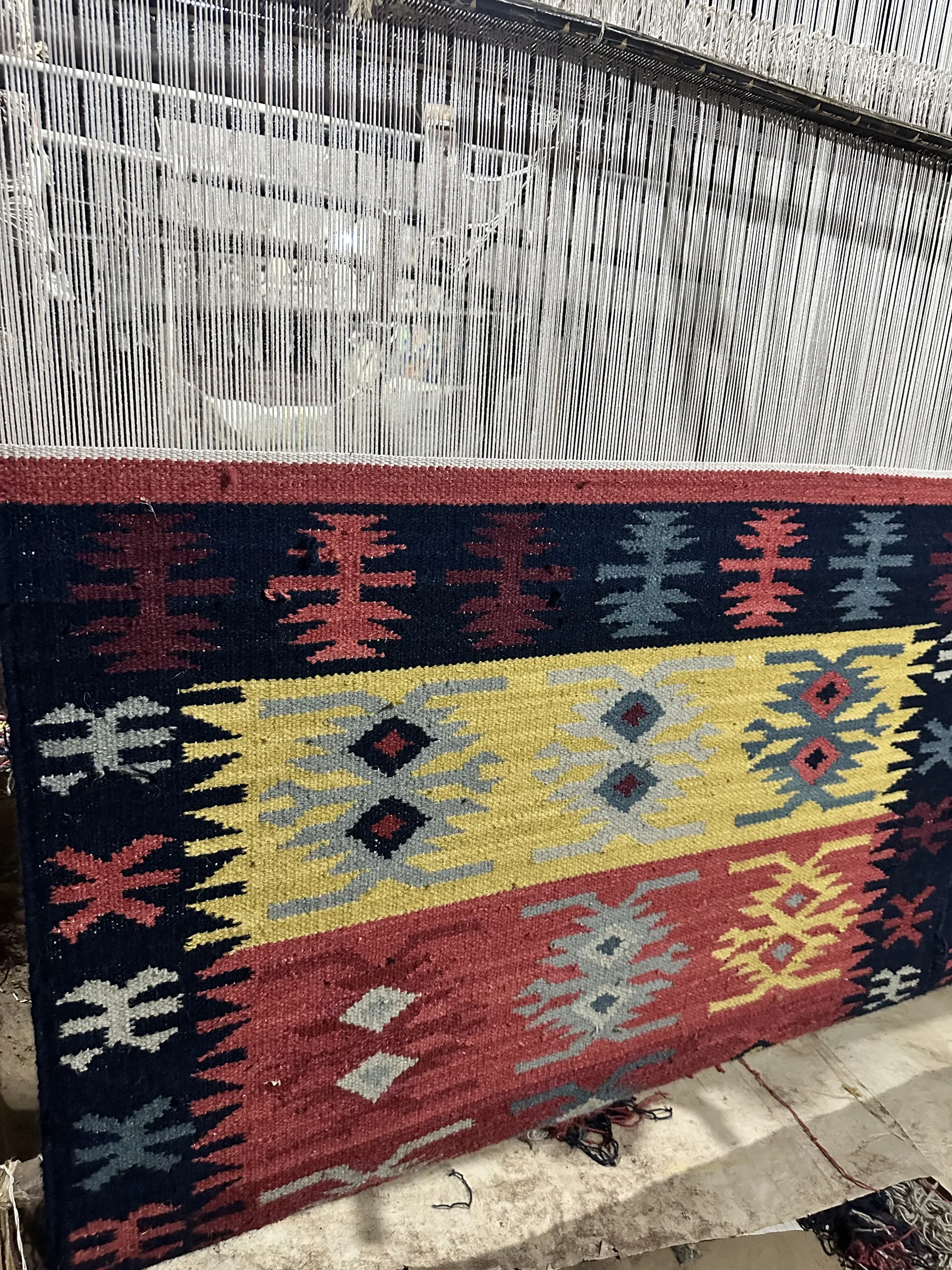 A geometric kilim in yellow, black, red and blue-grey on the vertical loom with warp threads above the finished band