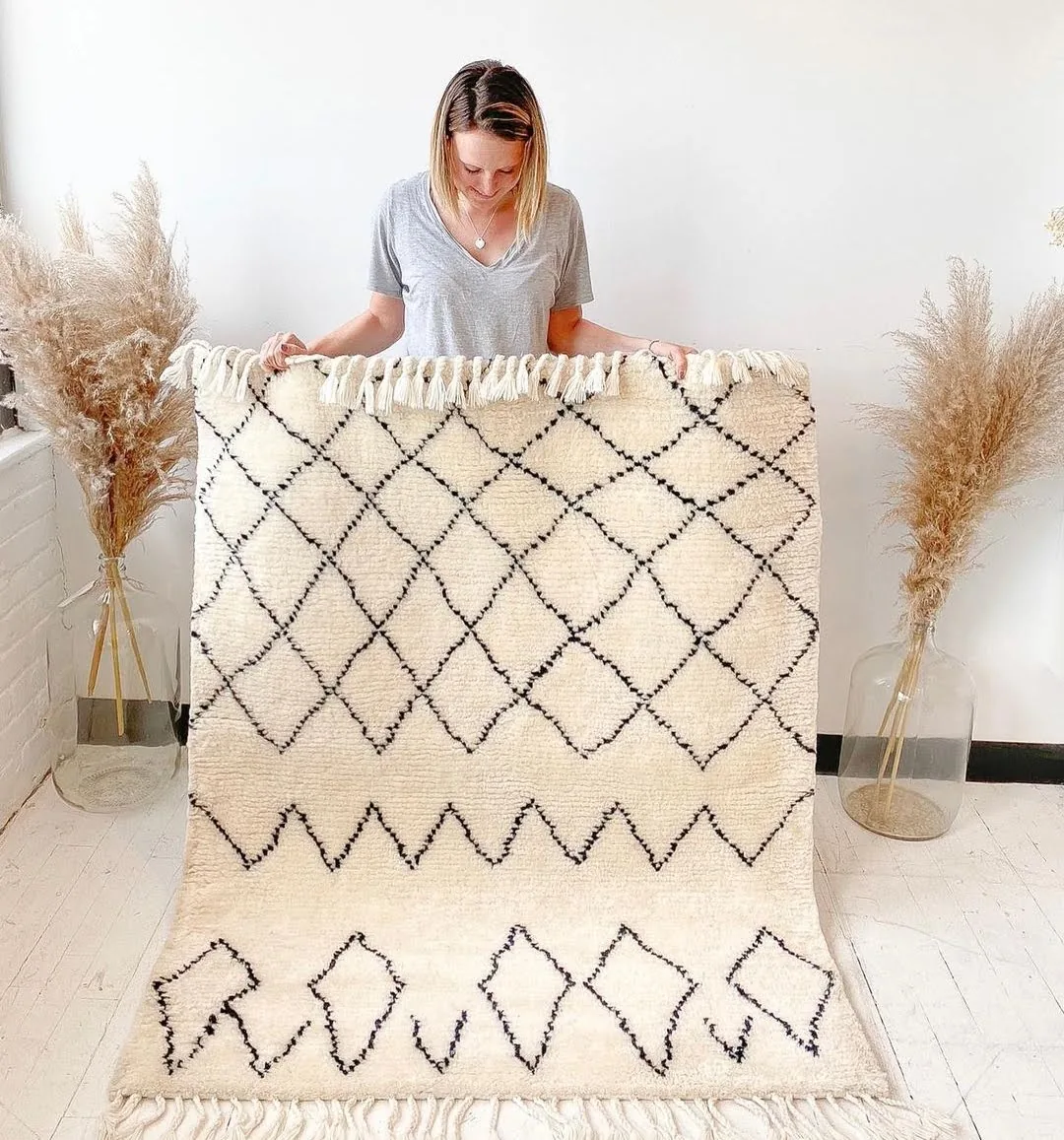 Berber-style hand-knotted wool rug in cream with a black diamond-lattice pattern and knotted tassel fringe, held up on a light interior wall between pampas grass and floor lamps