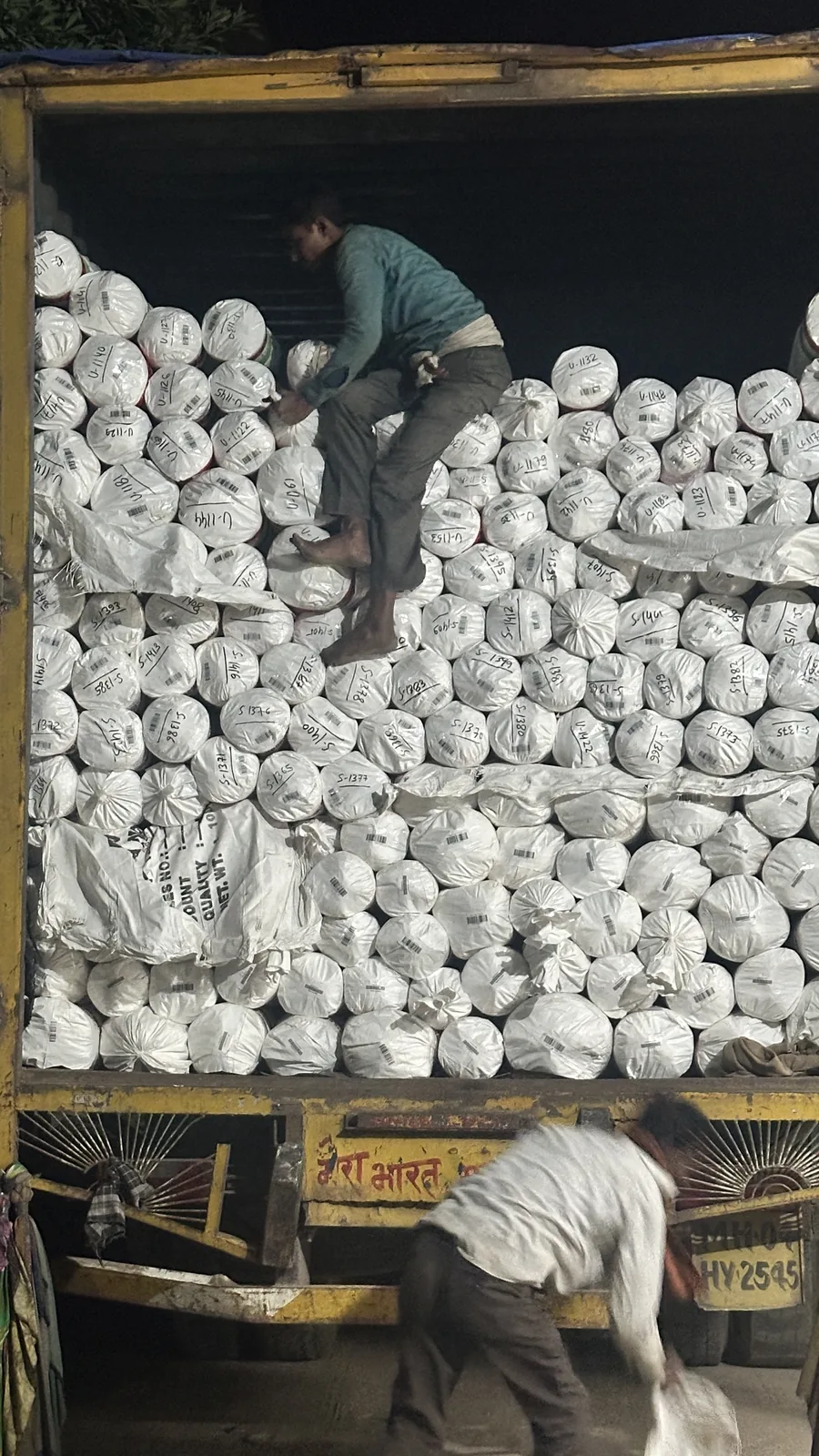 Wrapped rug bundles stacked to the roof of a truck bed at night, each roll labelled by SKU, one worker climbing the stack to place bundles and another closing the tailgate