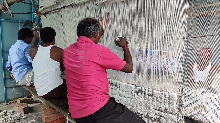Three weavers working side by side at a single wide loom